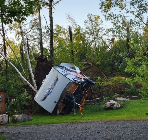 A large camping trailer is shown lying on its side, surrounded by trees that are uprooted and broken in half.