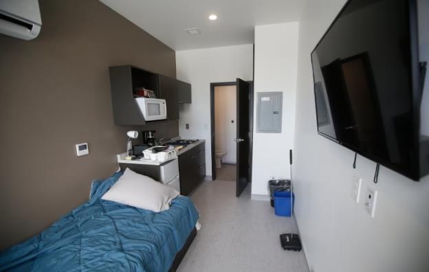 A small room holds a single bed with a blue blanket and pillow with a kitchenette along the left wall at the head of the bed and an open door at the end of the room showing a room with a toilet in it. A television is mounted on the right wall.