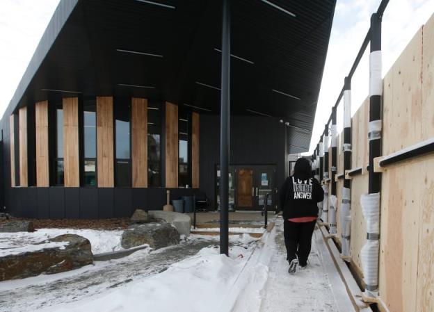 A woman walks down a snowy sidewalk beside a plywood wall towards a large black building with a ceiling jutting out and a circular wall of wood and glass.